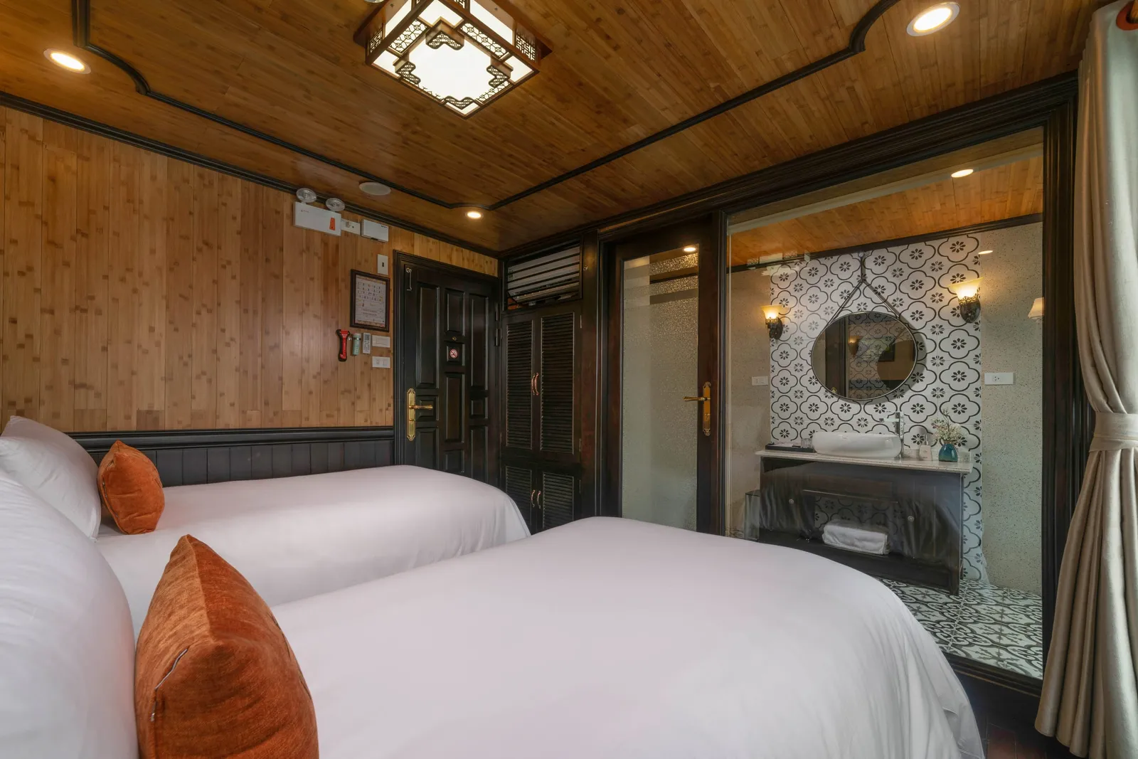 A twin-bed cabin with white linens, orange cushions, bamboo-paneled walls and ceiling, dark wood doors, and a glass-walled bathroom with patterned tiles and a round mirror.