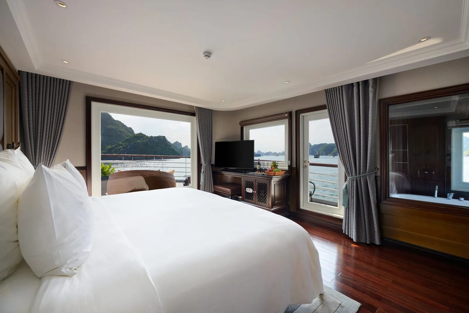A cruise cabin with a large white bed, dark wood floor and furniture, grey curtains, a TV and fruit basket, a glass-walled bathtub, and windows and a balcony door looking out onto a bay with limestone karst islands.