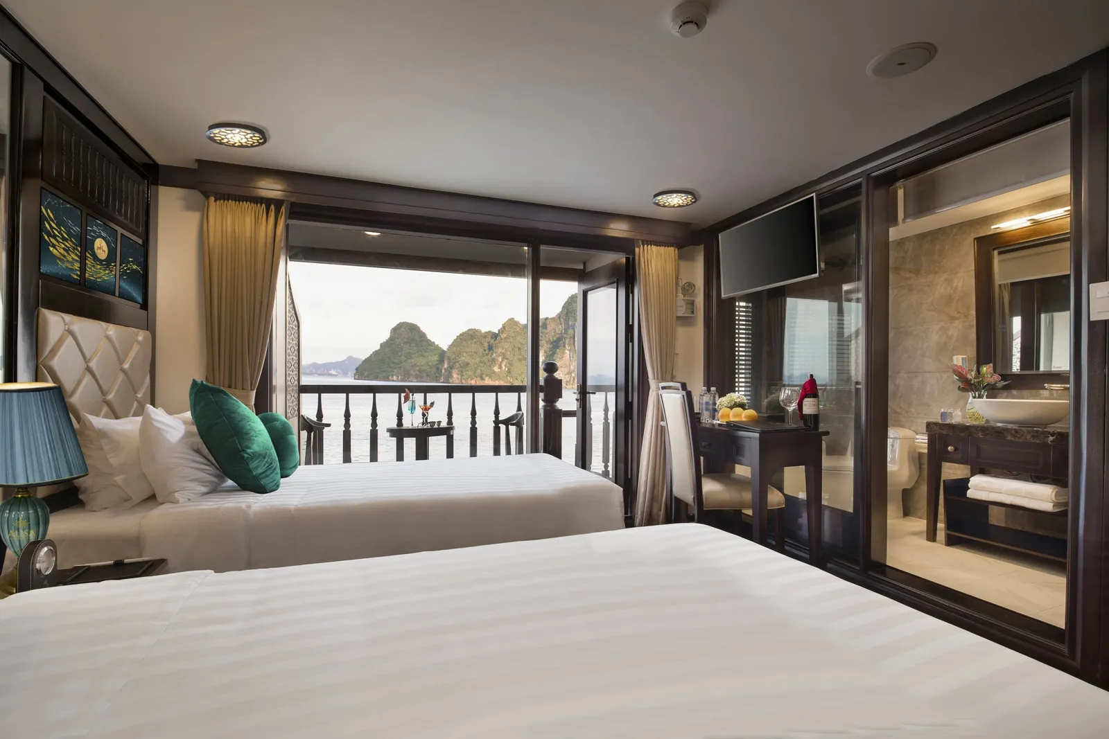 A twin-bed cruise cabin with white linens, green cushions, a desk with wine and fruit, an open glass bathroom, and a balcony door looking out to limestone karst islands on the water.