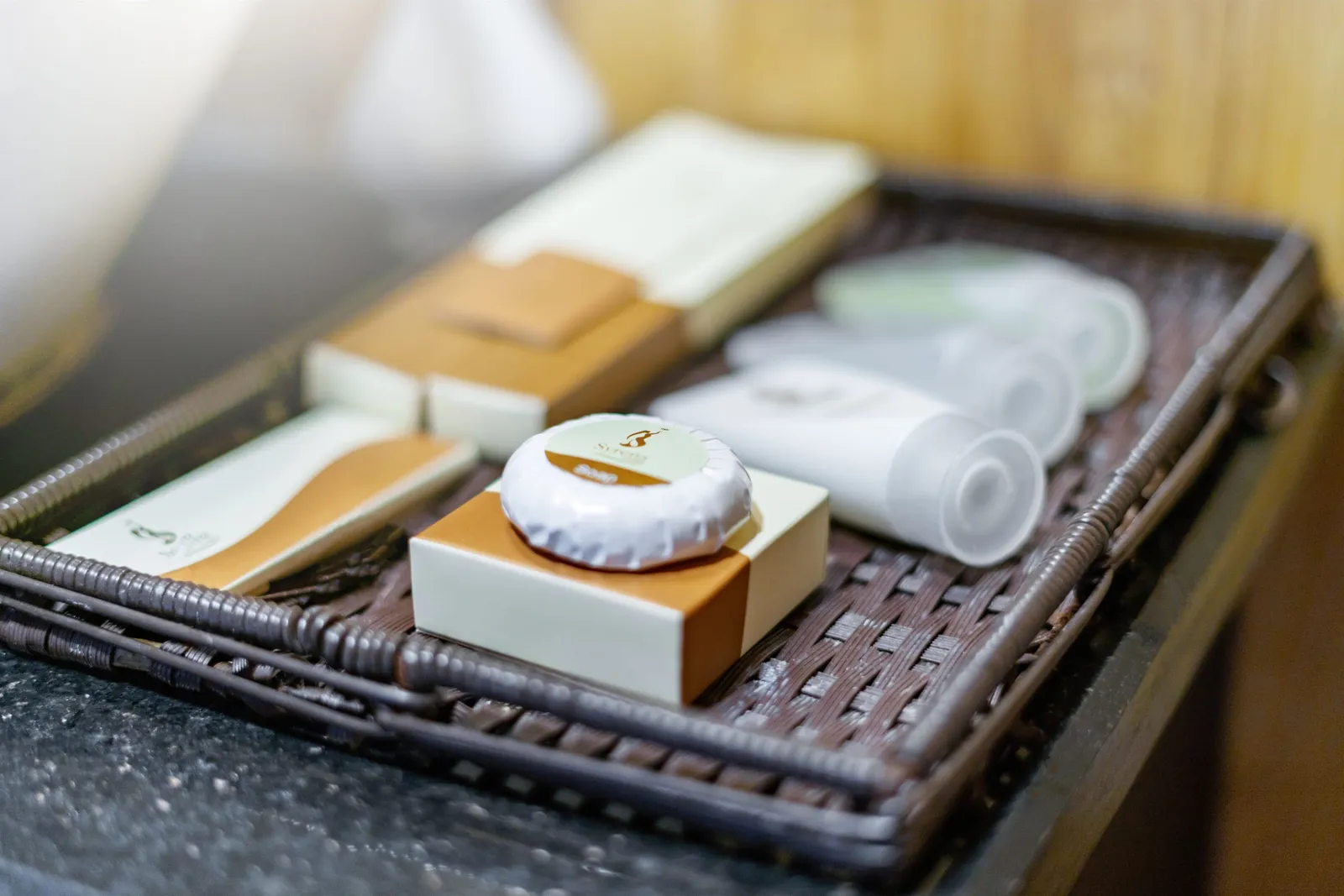 A woven wicker tray on a dark stone counter holds bathroom amenities: a wrapped soap bar on a box, small cartons and white tubes, with a small logo printed on the soap wrapper and packaging.