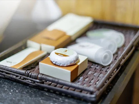 A woven wicker tray on a dark stone counter holds bathroom amenities: a wrapped soap bar on a box, small cartons and white tubes, with a small logo printed on the soap wrapper and packaging.