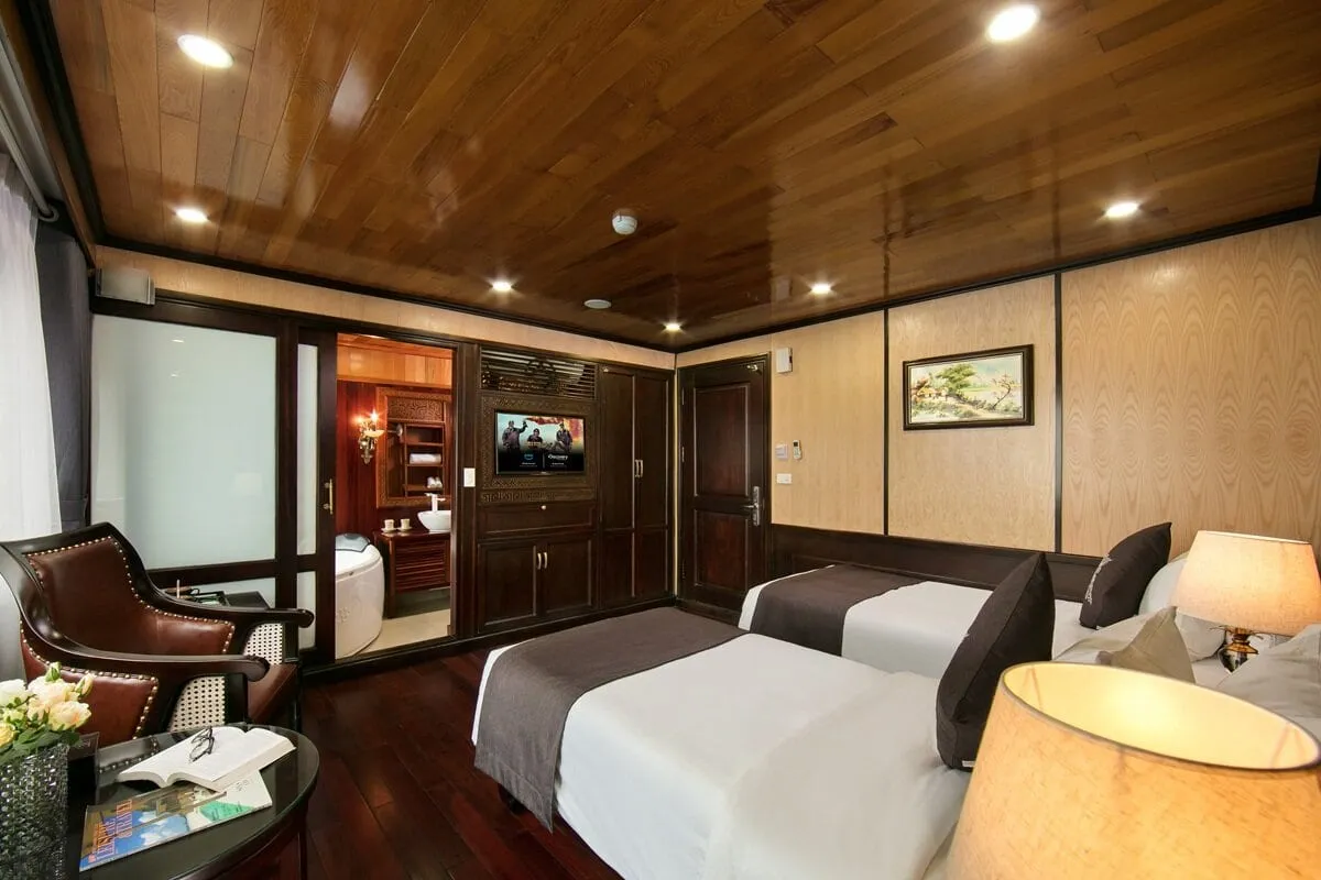 A wood-paneled cabin with two twin beds, a leather armchair, a wall-mounted TV, a framed landscape painting, and an open doorway to a bathroom with a round tub.