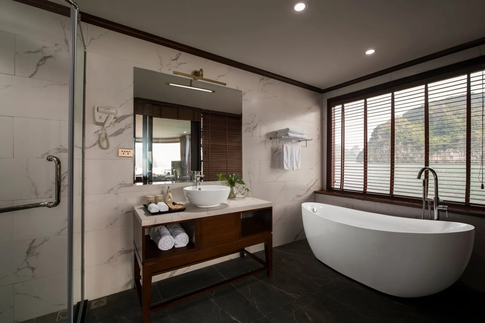 A marble-tiled bathroom with a freestanding white tub, wooden vanity with vessel sink, and a window with blinds showing a forested limestone karst and calm water outside.