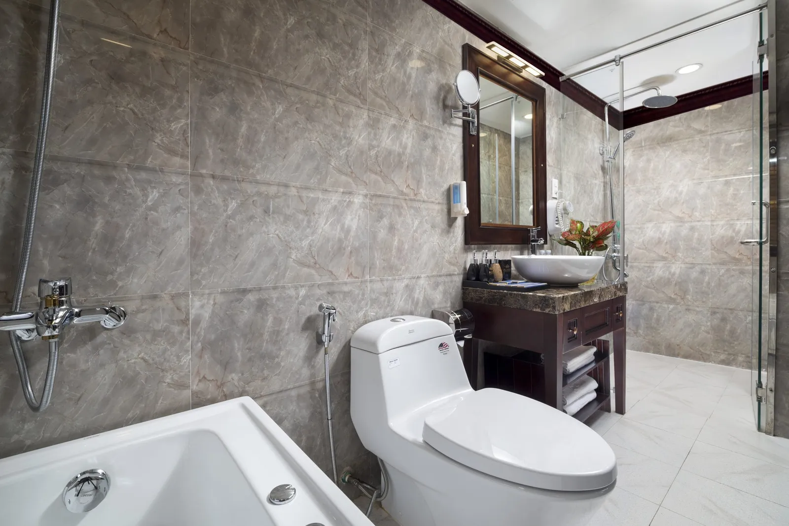 A marble-tiled en-suite bathroom with a bathtub, toilet, vessel sink on a dark wood vanity with mirror, and a glass-enclosed shower.