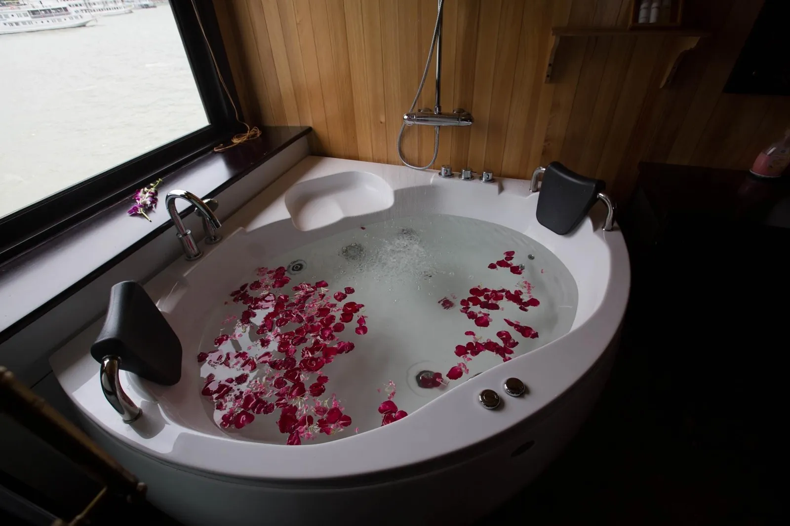 A white corner jacuzzi tub filled with water and red rose petals sits in a wood-paneled bathroom, with a window beside it showing calm water and a white boat in the distance.