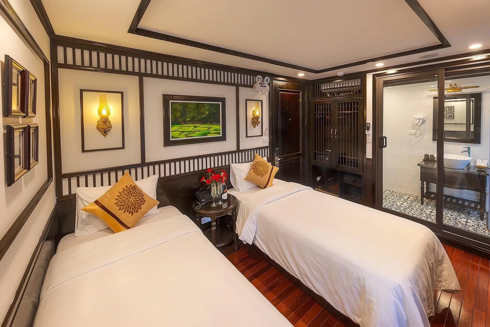 A twin-bed cabin with white linens, tan decorative cushions, dark wood lattice paneling, wall lamps, a green landscape painting, a small round table with red flowers and champagne glasses, and a glass-walled bathroom with a vessel sink.