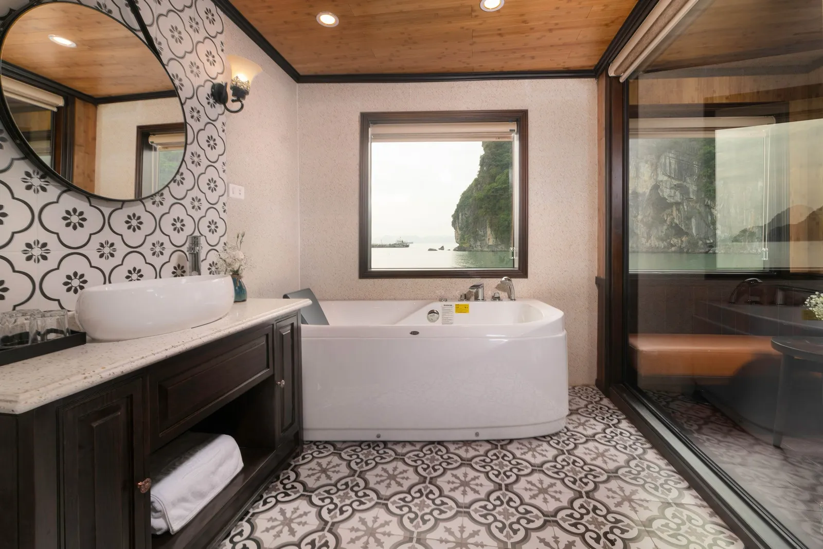 A cabin bathroom with a white corner bathtub, vessel sink on a dark wood vanity, patterned tiles, and a window and glass wall showing limestone karst cliffs over calm water.
