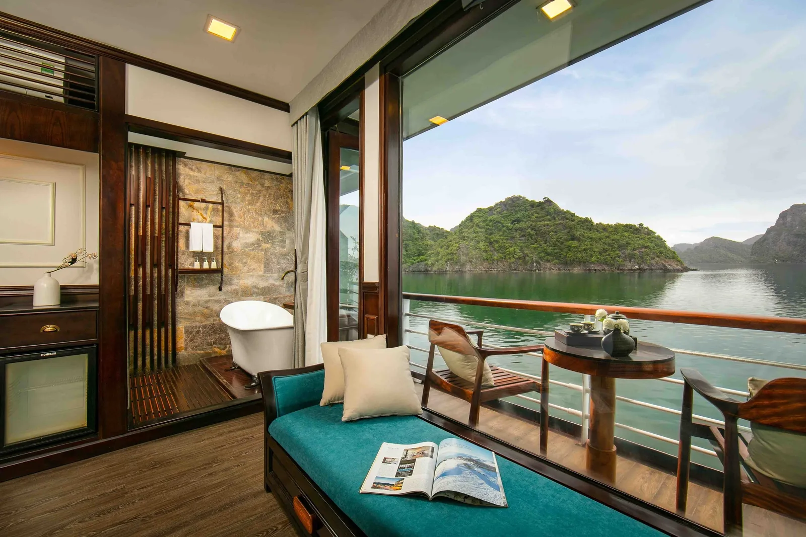 A wood-paneled cruise cabin with a teal daybed, freestanding bathtub and a private balcony with a small table and chairs, looking out over calm water toward green limestone islands.