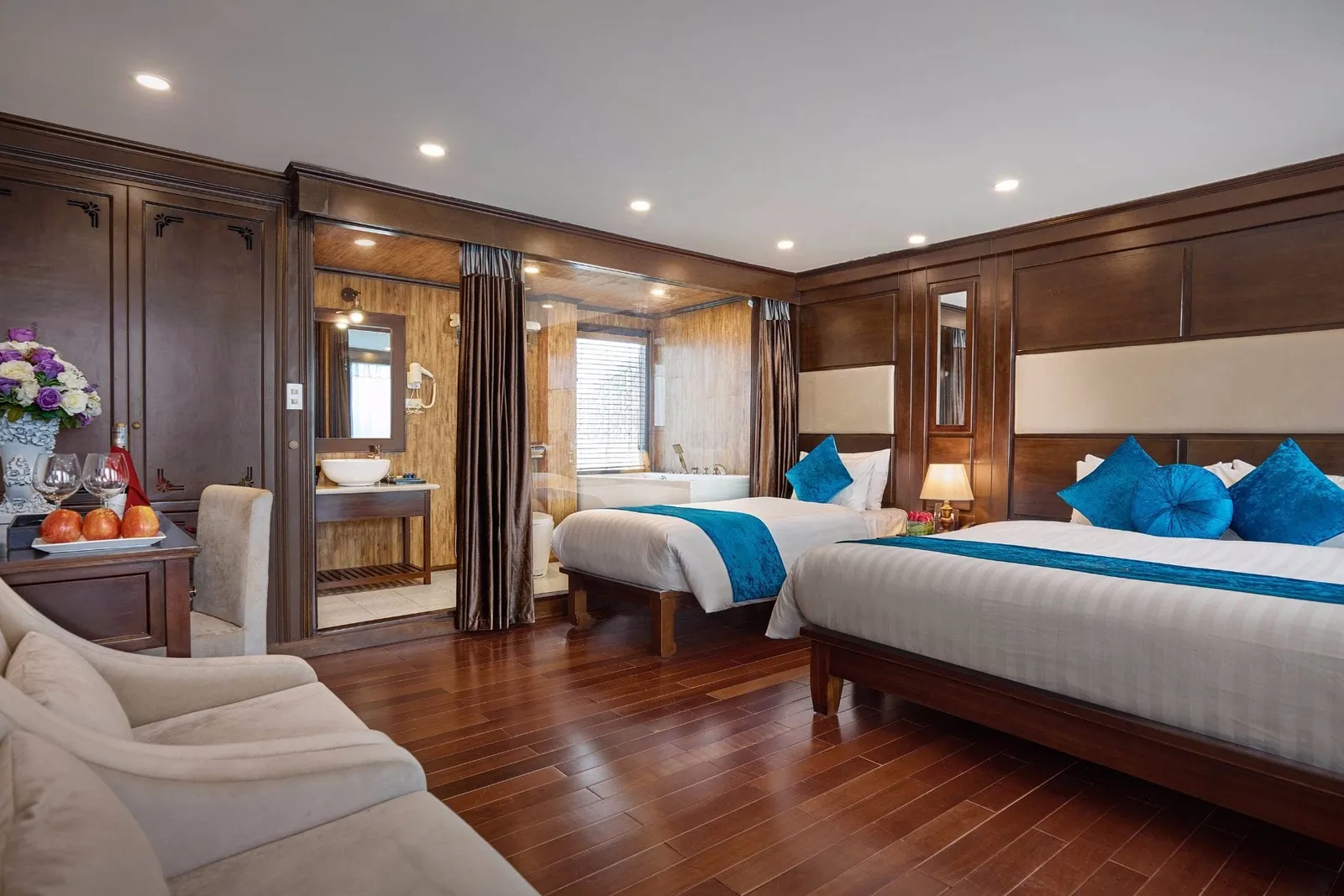 A wood-paneled cabin with a double bed and a single bed with teal accents, a glass-walled bathroom with a bathtub, a fruit plate and glasses on a desk, and a cream sofa on a hardwood floor.