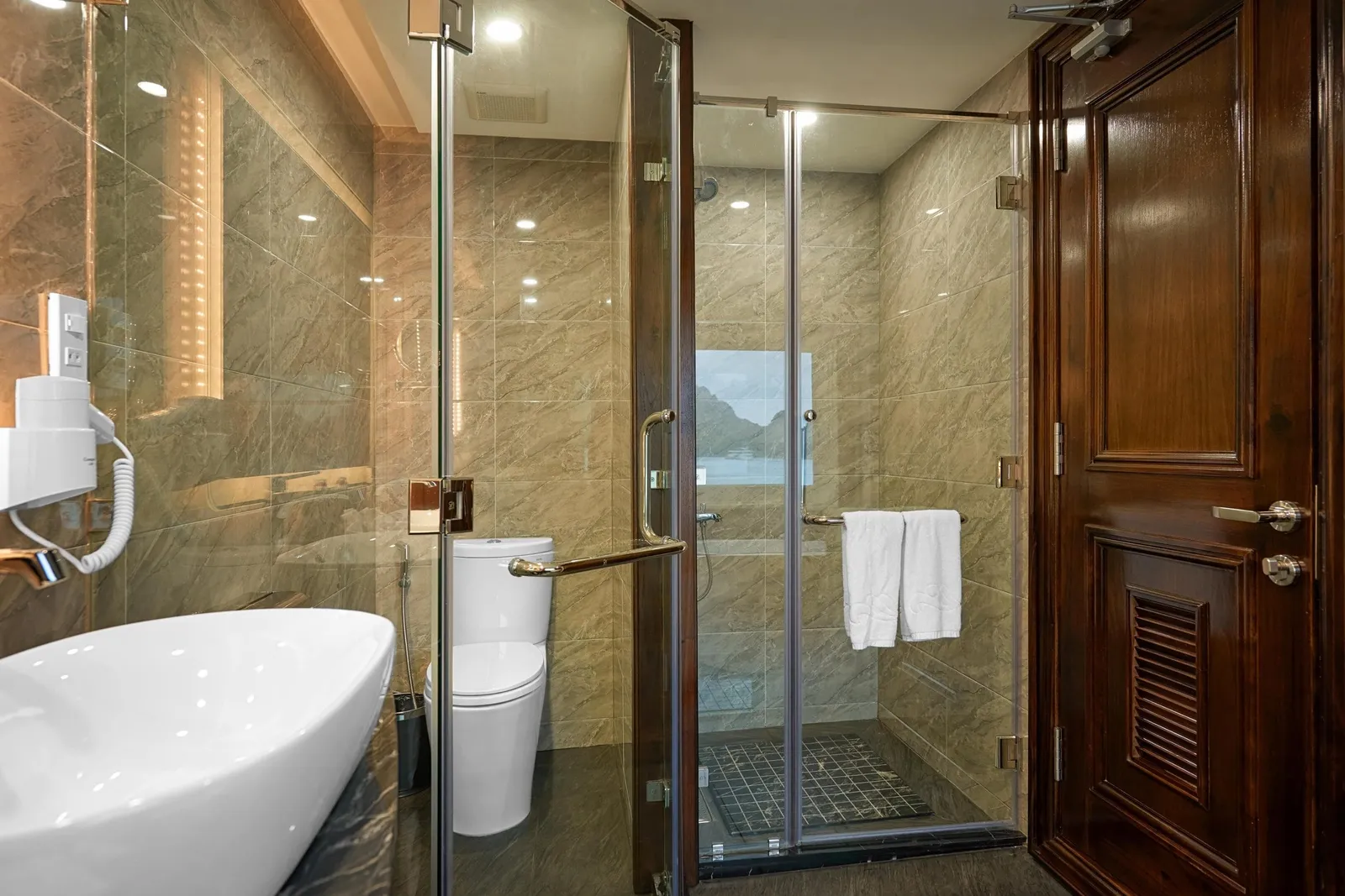 A marble-tiled en-suite bathroom with a white vessel sink, toilet, glass-enclosed shower with towels, and a dark wooden door, with a small window in the shower showing limestone karst and water.