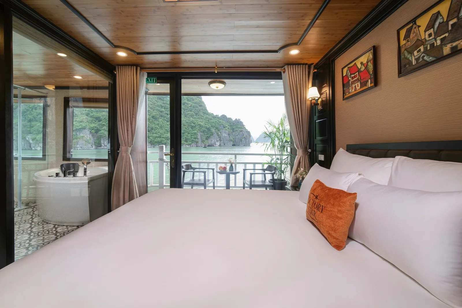 A cruise cabin with a white double bed and an orange embroidered 'Victory Star' cushion, a glass-walled bathtub on the left, and open balcony doors showing a railing, two chairs and limestone karst islands over green water.