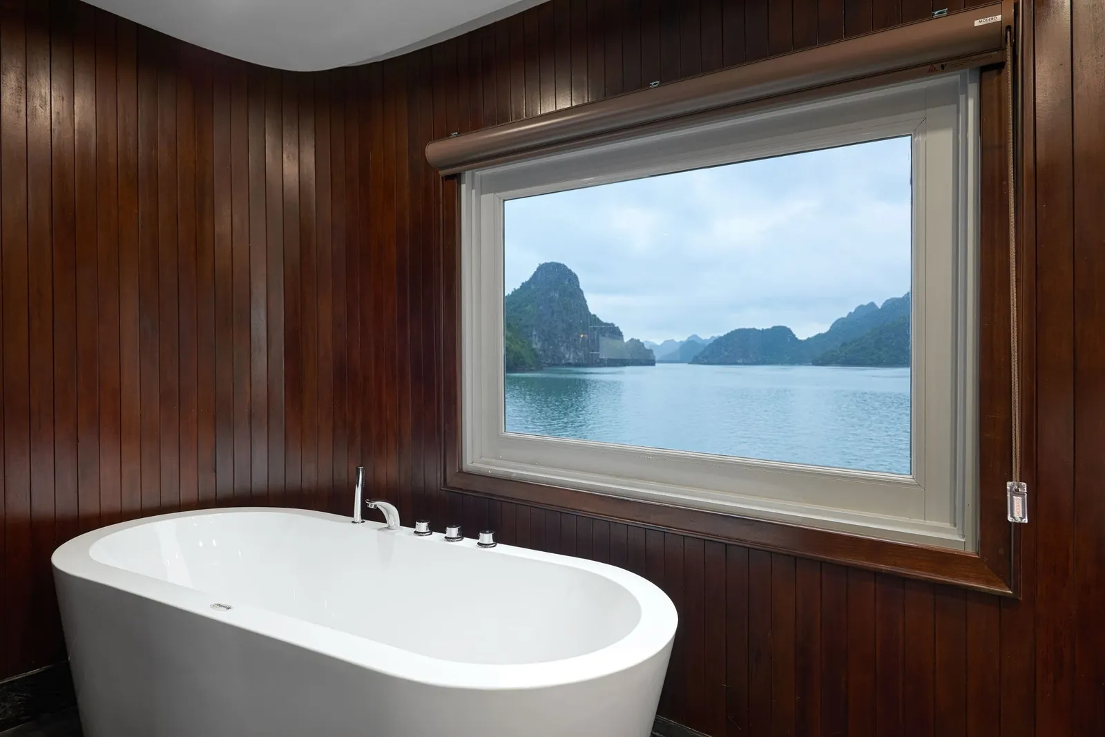 A white freestanding bathtub in a dark wood-paneled bathroom with a large window looking out onto calm water and limestone karst islands.
