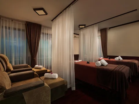 A dimly lit spa room with two massage beds with rolled white towels, sheer curtains dividing the space, and two brown upholstered footrest recliners in the foreground.