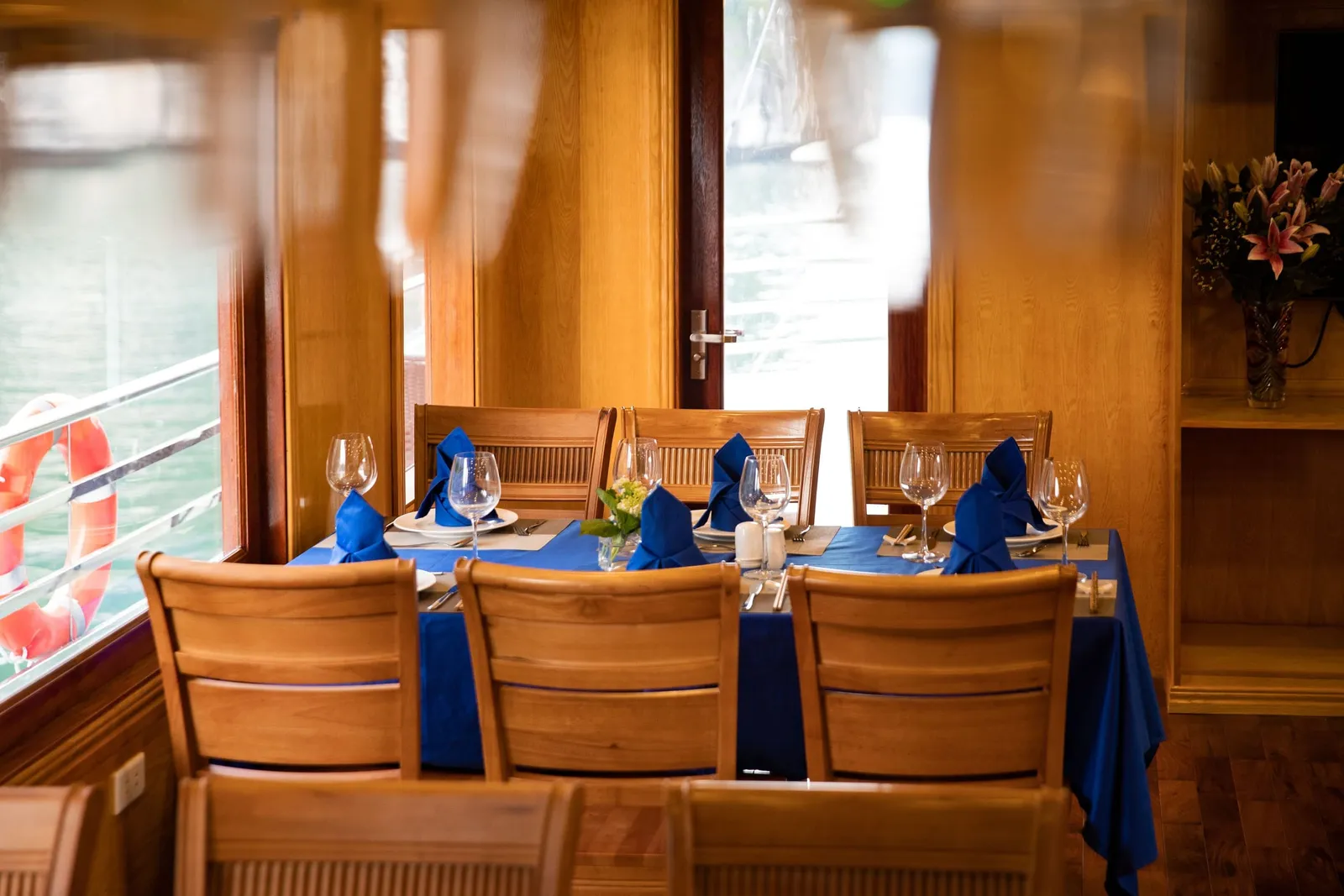 A wood-paneled boat dining room with a table set in blue cloth and napkins, wine glasses and wooden chairs, with a life ring outside the window and a flower vase on a shelf at right.