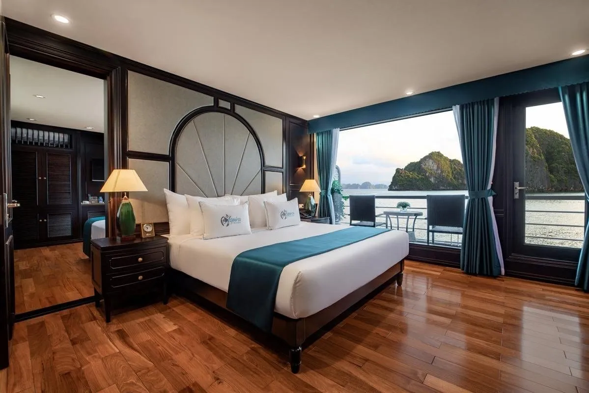A cruise cabin with a large white bed with teal runner and logo-embroidered cushions, dark wood furniture, a wooden floor, and glass doors opening to a balcony overlooking calm water and limestone karst islands.