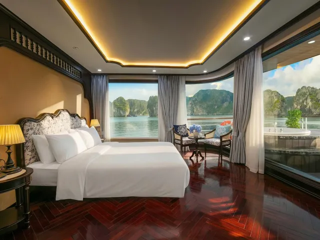 A cruise cabin with a white double bed, dark herringbone wood floor, and floor-to-ceiling windows overlooking calm water and limestone karst islands, with a private jacuzzi on the balcony.