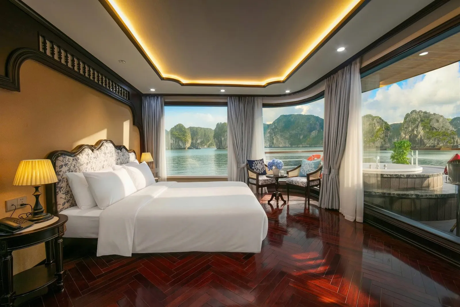 A cruise cabin with a white double bed, dark herringbone wood floor, and floor-to-ceiling windows overlooking calm water and limestone karst islands, with a private jacuzzi on the balcony.