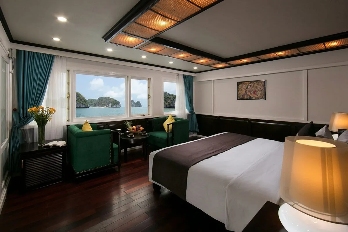 A cabin with a bed, two green armchairs, a fruit bowl and flowers, with windows showing limestone karst islands in calm green water.