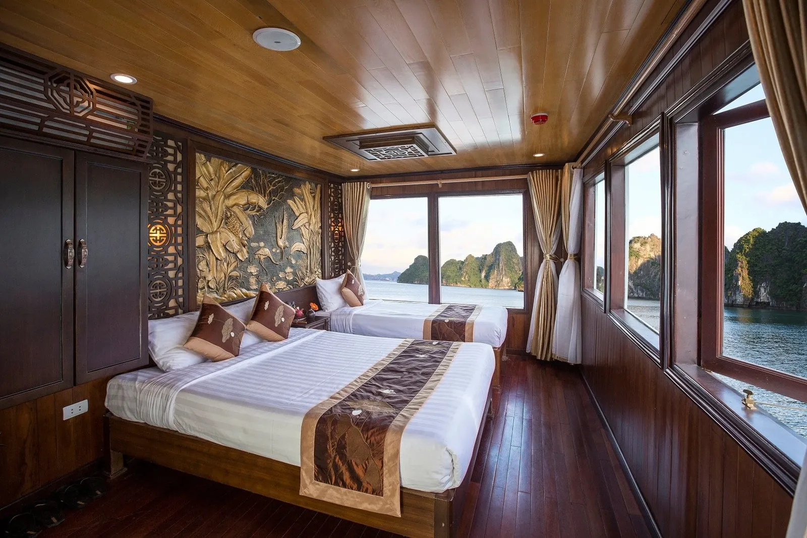 A wood-paneled cruise cabin with two beds with brown runners and cushions, a carved golden wall relief, and large windows showing limestone karst islands on calm water.