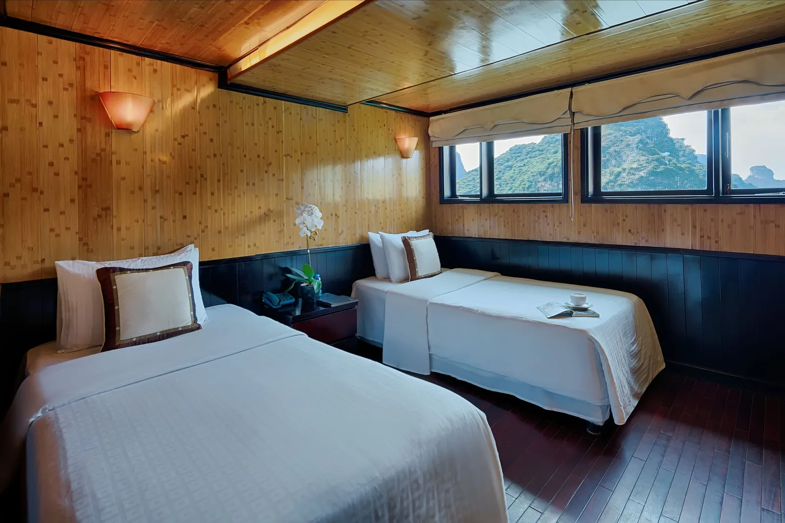 A wood-paneled cabin with two twin beds in white linens, a bedside table with an orchid and phone, and windows showing green limestone karst hills.