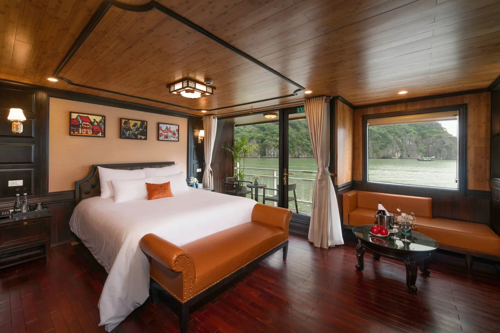 A wood-paneled cruise cabin with a white-linen double bed, tan leather bench and sofa, and a glass door and window opening onto a balcony and a green bay with limestone karst cliffs.