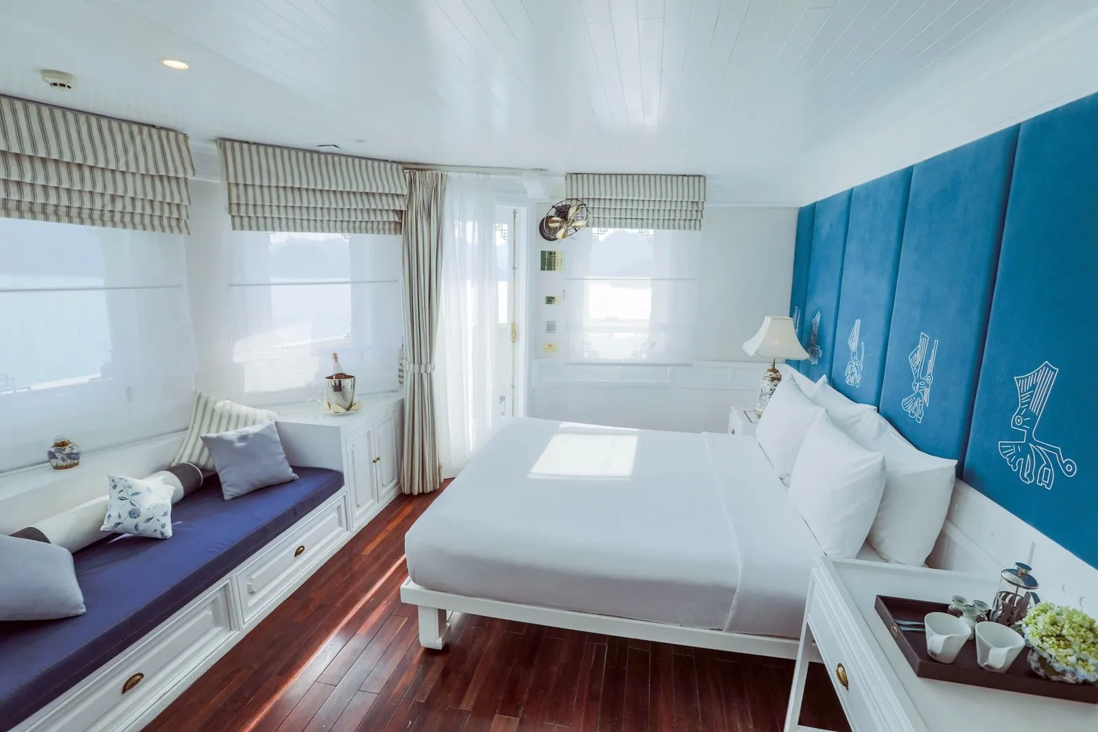 A bright cabin with a white double bed, blue padded headboard panels with white line motifs, a navy window seat with cushions, striped roman blinds, and a wooden floor, with hazy water visible through the windows.