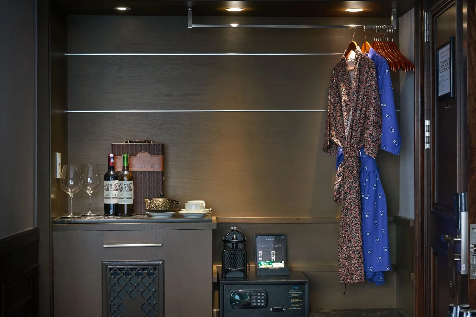 A cabin alcove with a counter holding two wine glasses, two wine bottles, a gift box, a teapot and cup, a coffee machine and a room safe below, with two robes hanging on wooden hangers beside an open door.