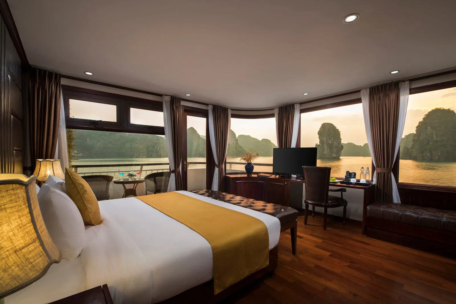 A cruise cabin with a double bed, gold runner, wood floor, desk with TV and panoramic windows looking out on limestone karsts in a bay at sunset.