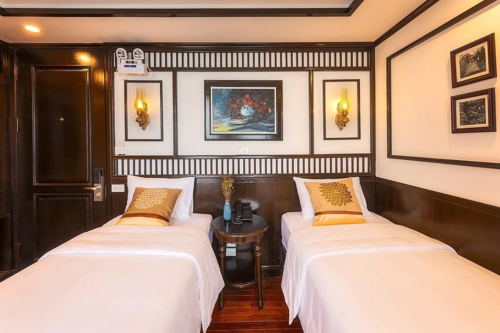 A twin-bed cabin with white linens, gold patterned cushions, dark wood paneling, lantern-style wall lamps, a framed floral painting, and a round bedside table with a phone and vase.