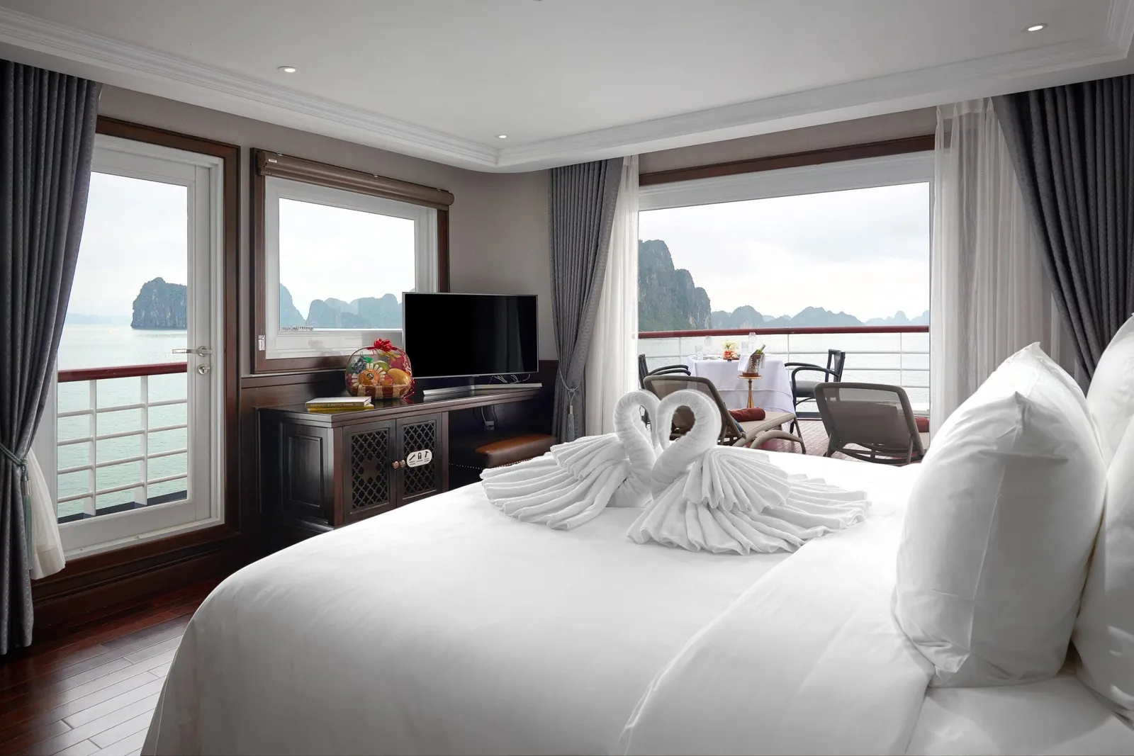 A cruise cabin with a white bed topped with towel swans, a TV and fruit basket, and windows and a balcony with a dining table overlooking limestone karsts on the water.