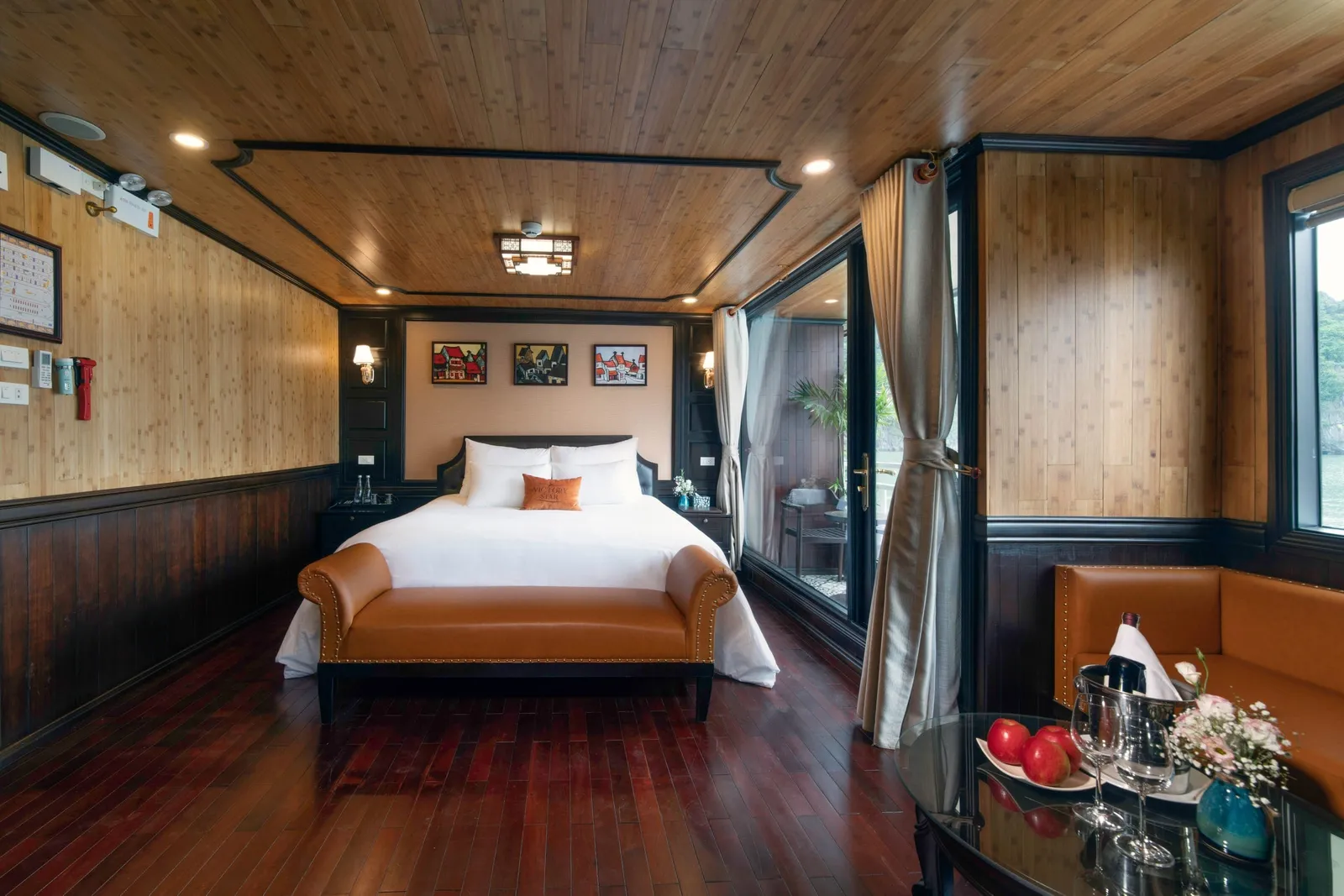 A wood-paneled cruise cabin with a white double bed, tan leather bench, glass table with fruit, champagne bucket and flowers, and a balcony door opening toward water and a green karst hillside.