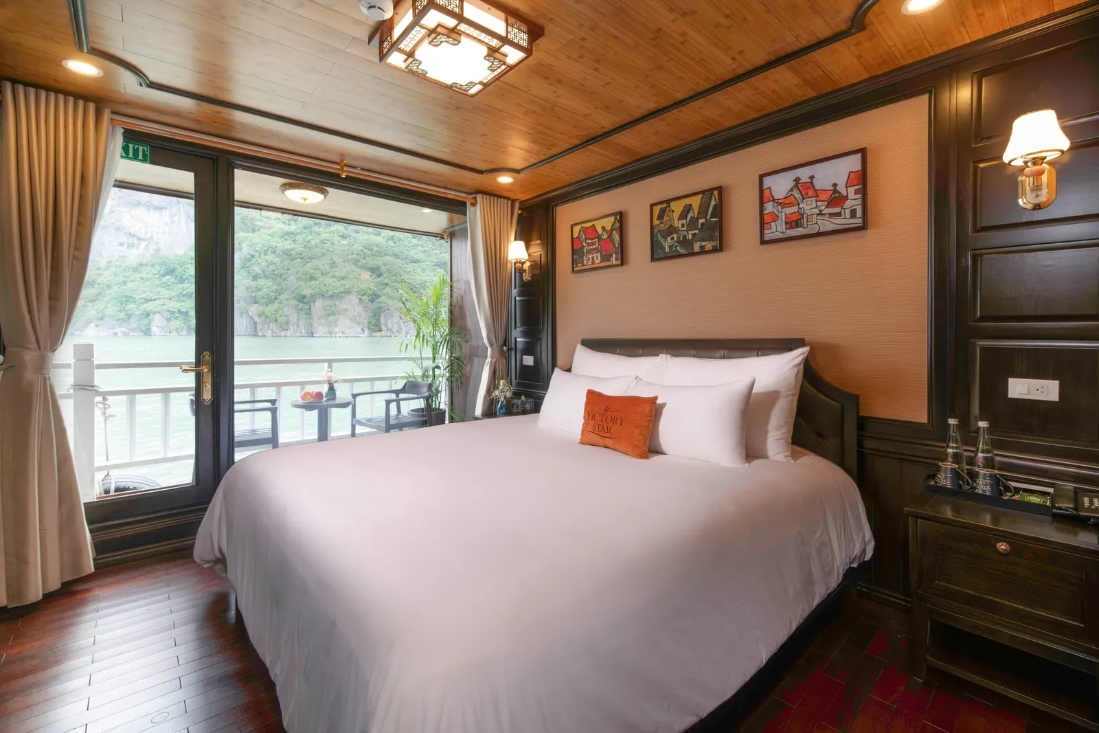 A wood-paneled cruise cabin with a white double bed and an orange 'Victory Star' embroidered cushion, with glass doors opening onto a balcony that overlooks green karst cliffs and calm water.