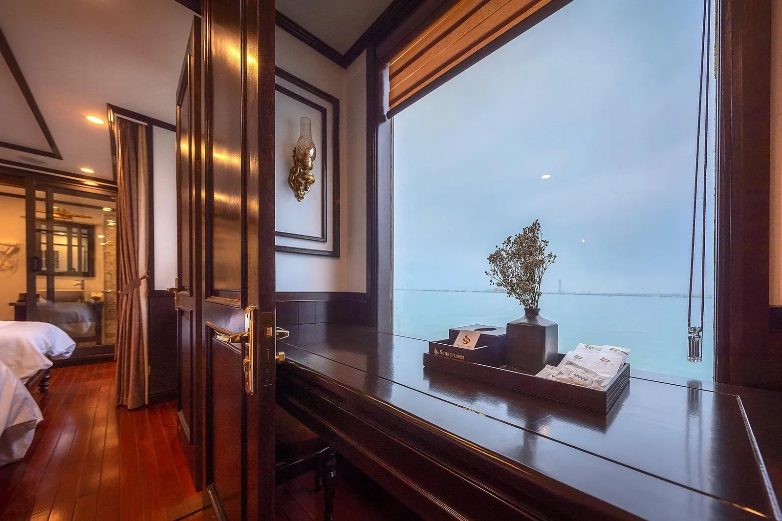 A dark wood-paneled cabin with a desk by a large window holding a tray with a 'Seacruises' logo, a vase of dried flowers and tissues, overlooking calm hazy water, with a bed visible at left.