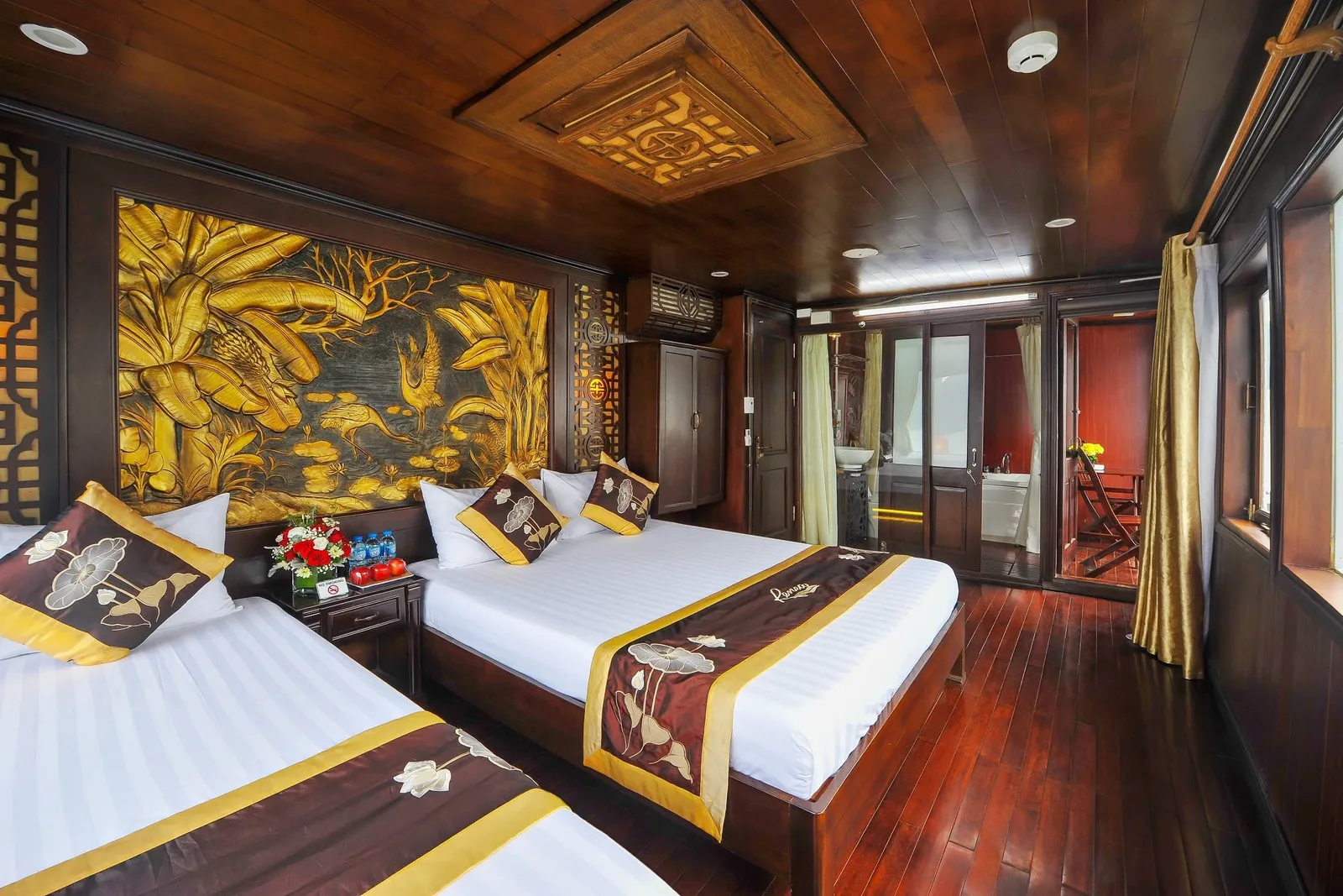 A dark-wood cabin with two twin beds in white linens with gold and brown lotus-pattern runners, a carved golden relief panel on the wall, a bedside table with flowers and water bottles, and a glass-doored bathroom and balcony beyond.