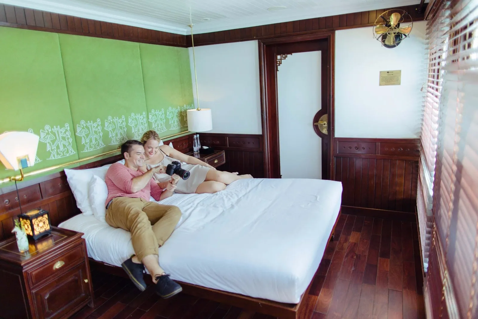 A smiling man and woman lie on a white bed in a dark-wood cabin with a green padded wall panel, looking at a camera the man holds, with a small plaque and a brass fan on the wall and window blinds on the right.