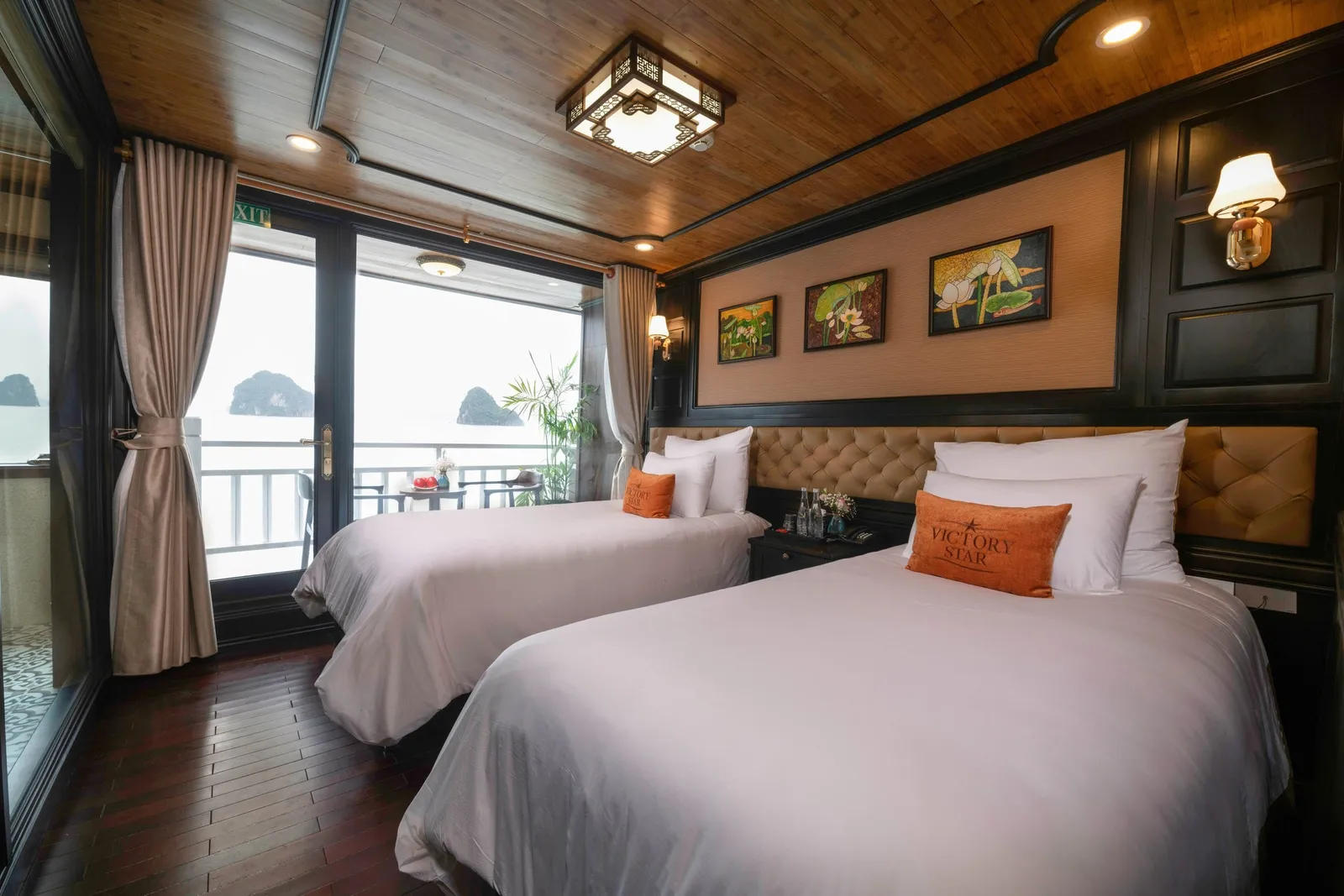 A wood-paneled cruise cabin with two twin beds, an orange cushion embroidered 'Victory Star', and glass doors opening to a balcony overlooking misty limestone karsts.