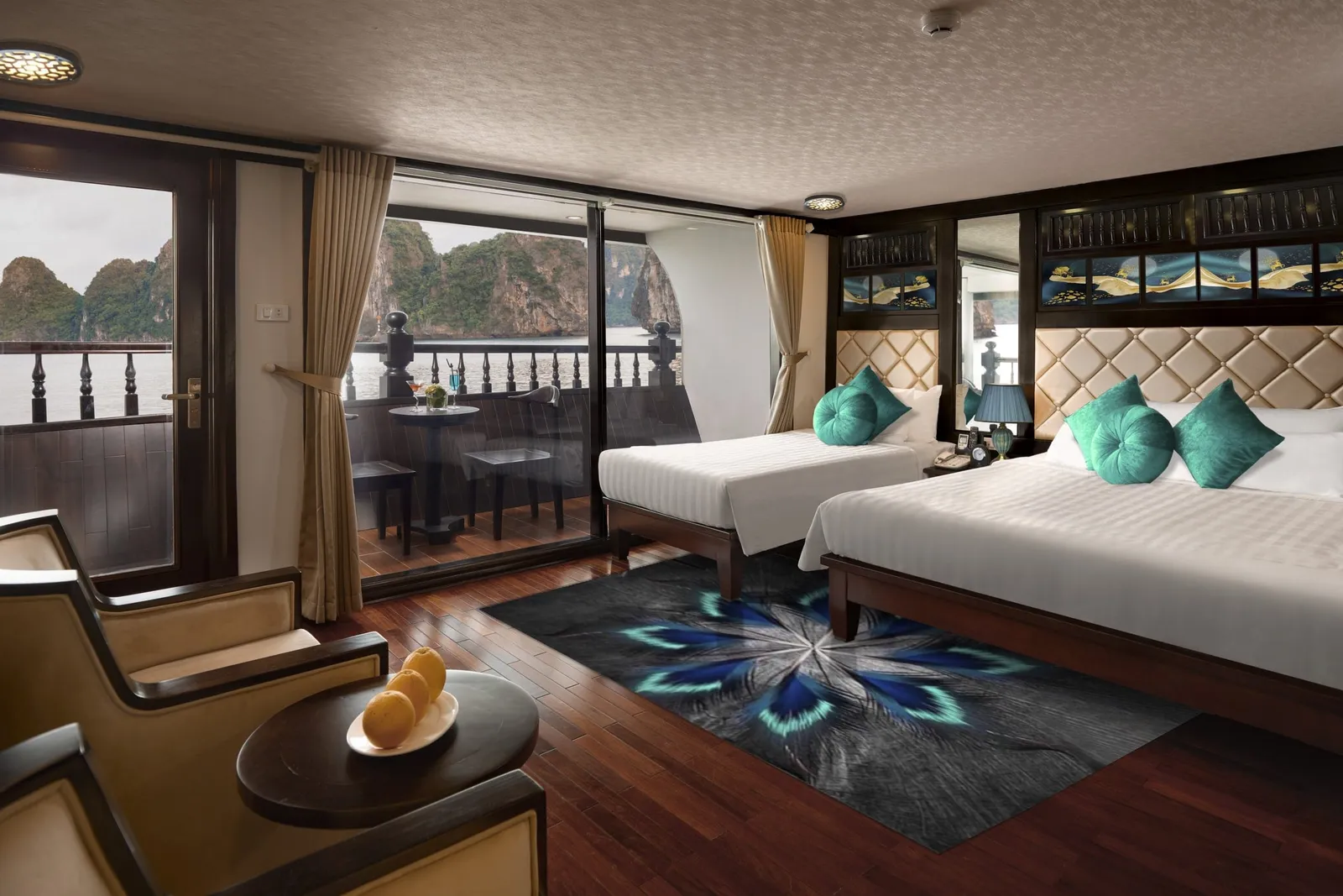 A cruise cabin with two beds with teal cushions, a peacock-feather rug, armchairs and a fruit plate, with glass doors opening to a balcony overlooking limestone karst islands on the water.