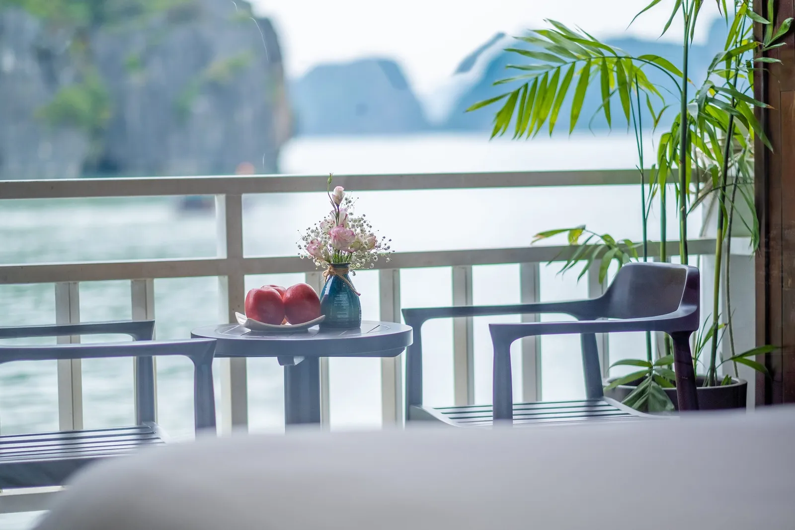 A small balcony table with a plate of red apples and a vase of pink flowers, two chairs, a potted bamboo plant, and a white railing overlooking calm water and misty limestone karsts.