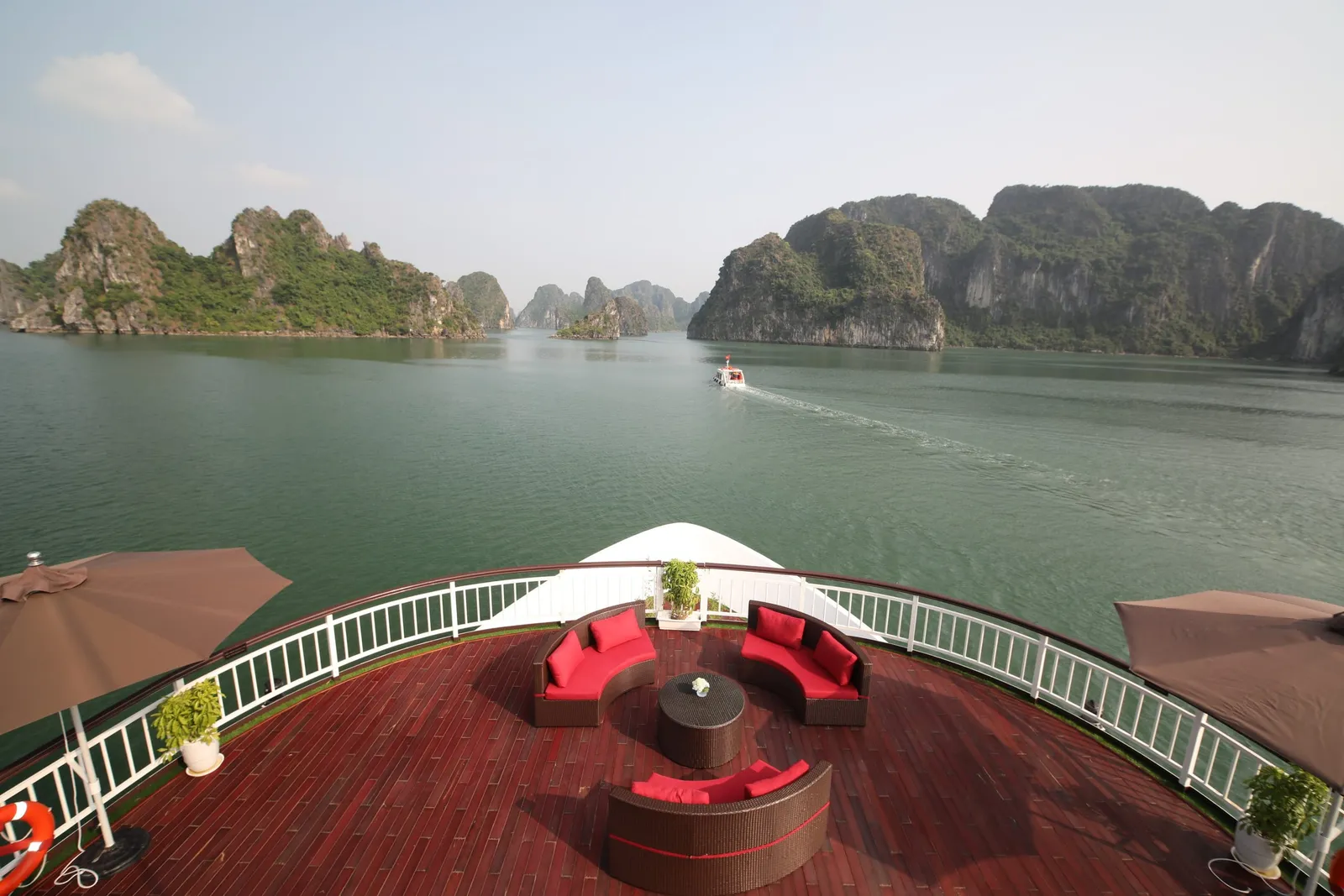 A red-brown wooden upper deck with wicker sofas with red cushions, a round table, brown umbrellas and a white railing, overlooking green water and limestone karst islands with a small boat in the distance.