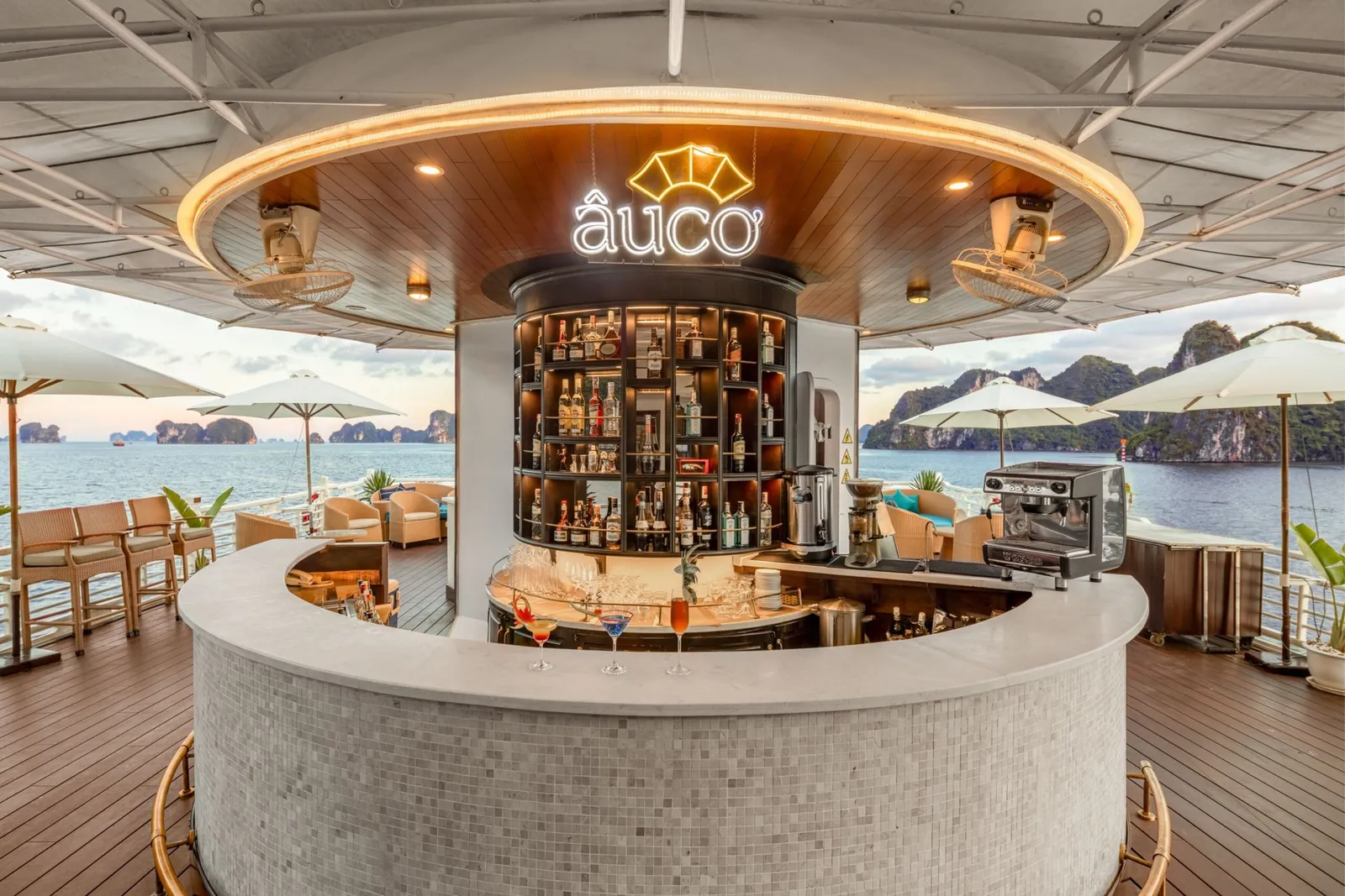 A circular open-air bar on a cruise ship's upper deck with an illuminated 'âuco' neon sign, cocktails on the counter, umbrellas and chairs around, and limestone karst islands over the water at dusk.
