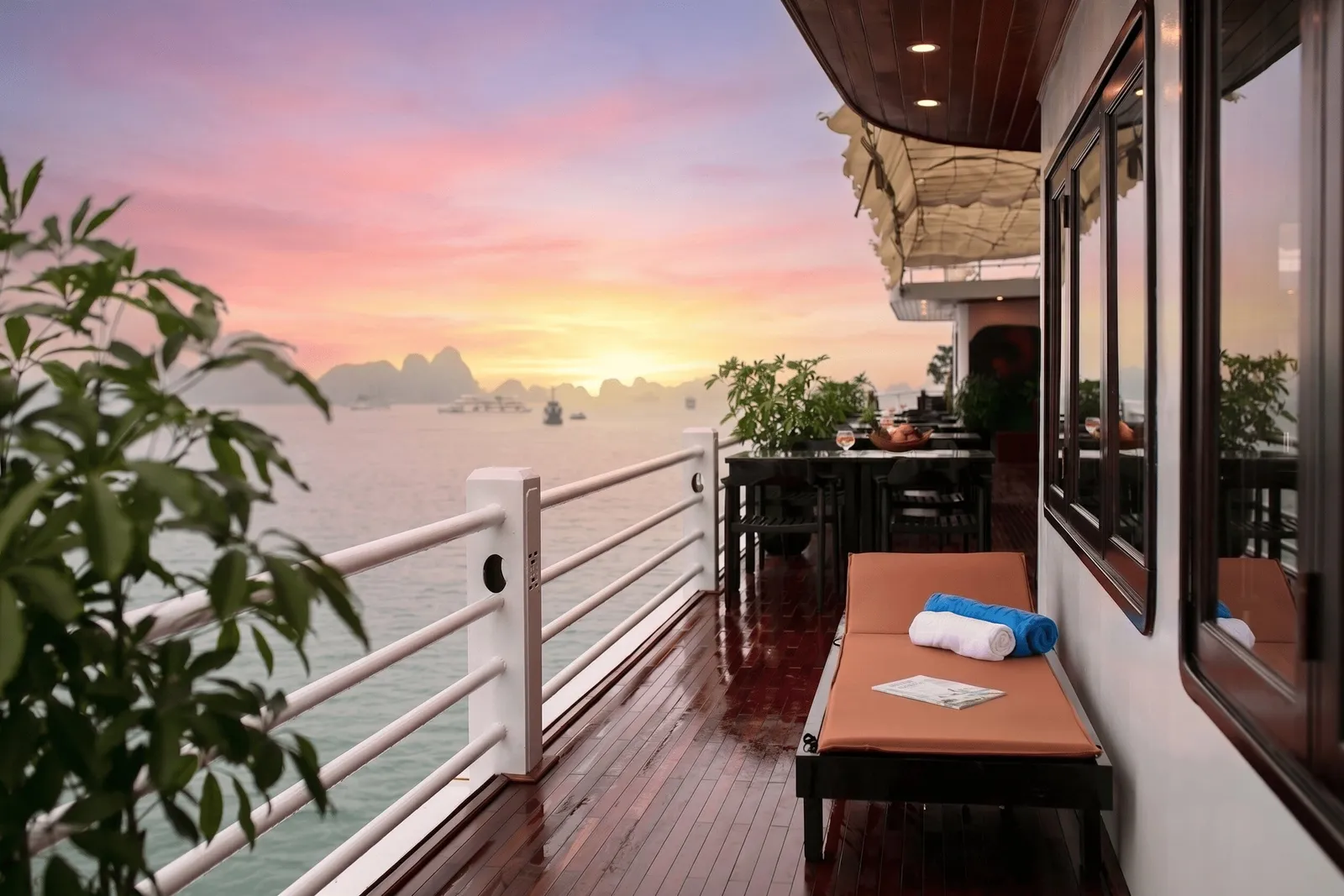 A wooden side deck of a cruise boat at sunset with a white railing, a cushioned lounger with rolled towels, a dining table with plants, and limestone karsts on the calm bay in the distance.