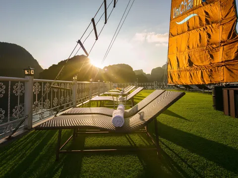 Sun deck with artificial grass, wicker loungers with rolled towels, an ornate white railing and an orange sail printed with 'UniCharm' logo, with limestone karsts silhouetted against a low sun.