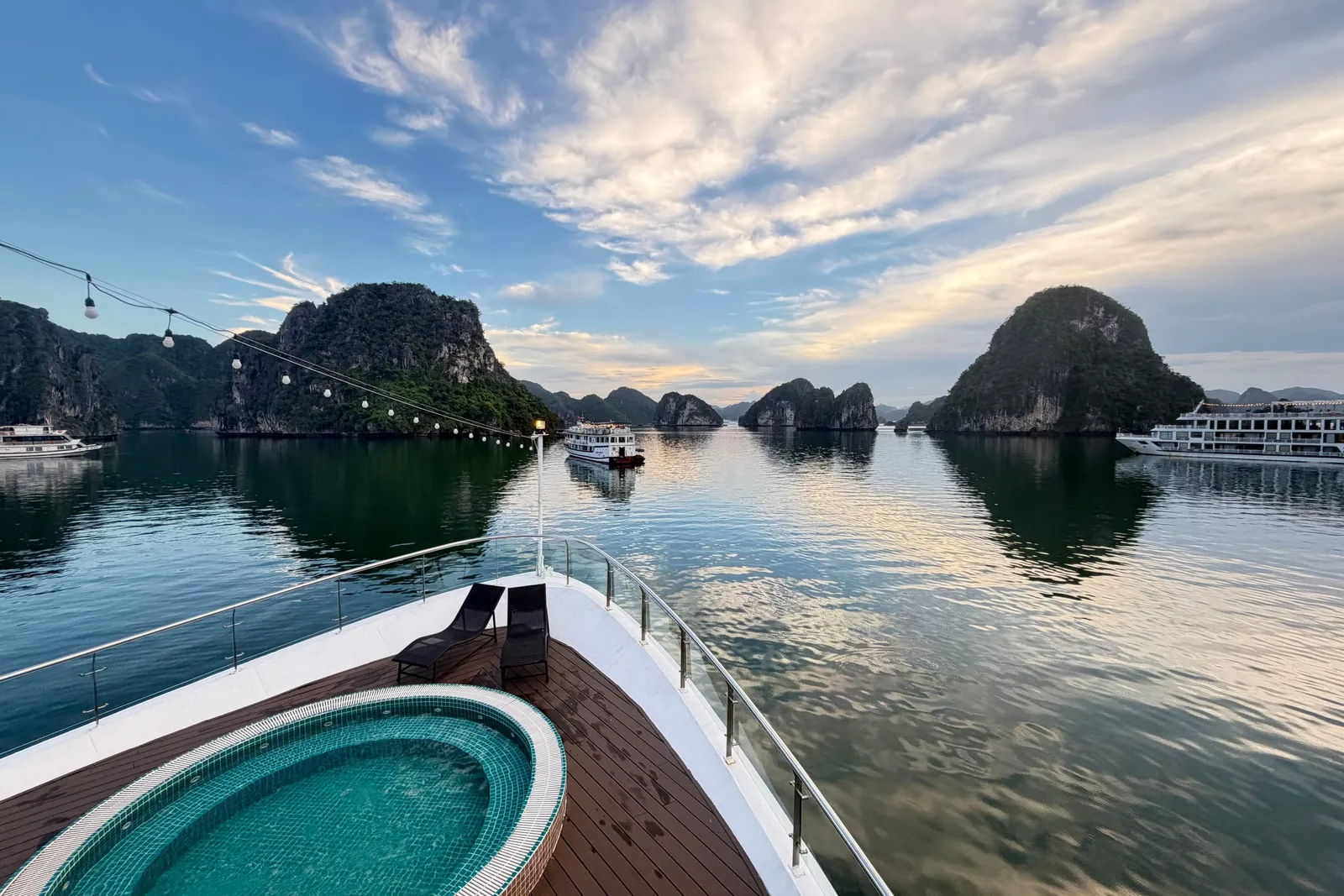 The bow deck of a cruise boat with a round tiled jacuzzi, two black loungers and string lights, facing calm water, limestone karst islands and other boats under a cloudy blue sky.