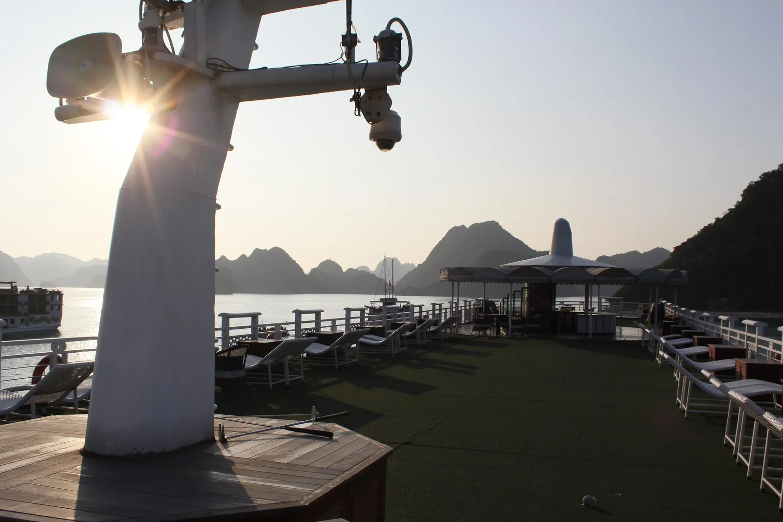 Sun-lit sundeck of a cruise boat with lounge chairs, a shaded bar area and a mast with a camera, looking out over calm water toward limestone karst hills and another boat.