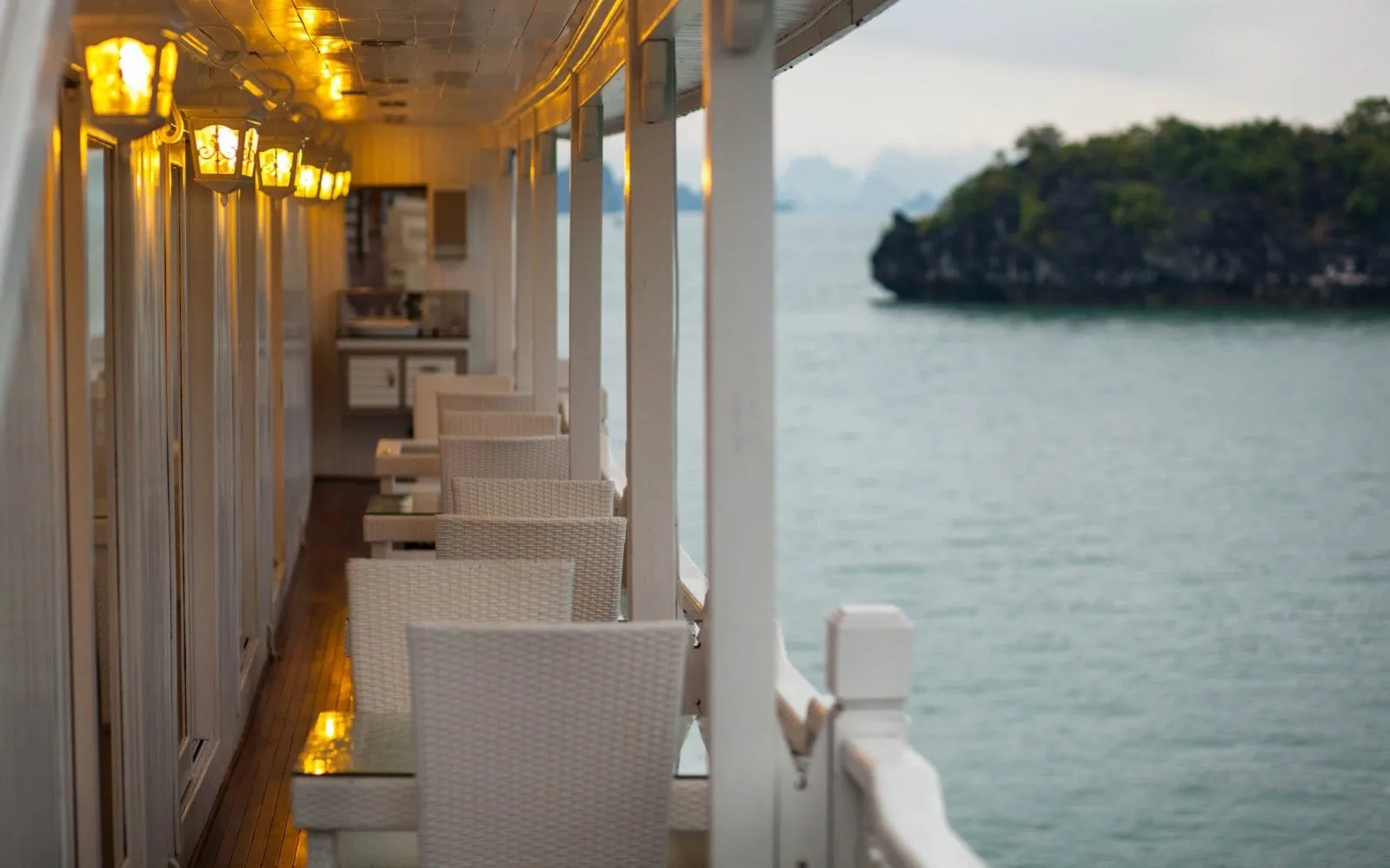 A covered side deck of a boat with white wicker chairs and tables, lit wall lanterns, and a wooden floor, looking out over calm water toward a small tree-covered rocky island and hazy karst silhouettes.