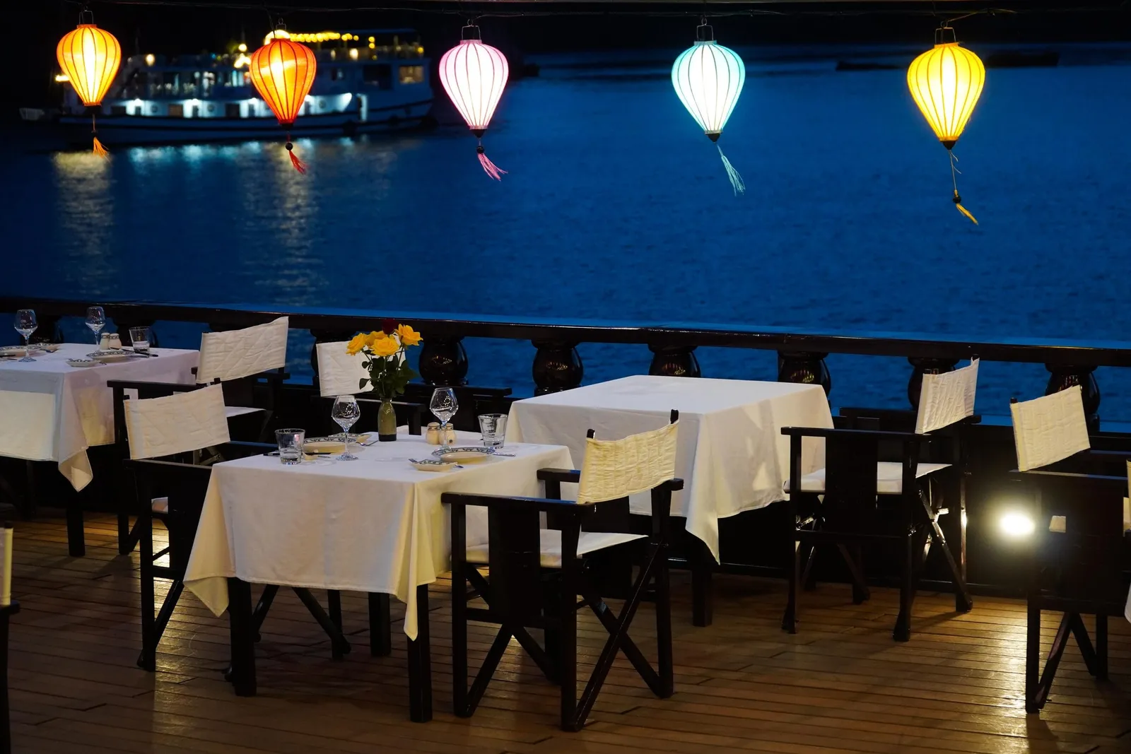 Dusk view of a wooden boat deck set with white-clothclad dining tables and folding chairs under glowing silk lanterns, overlooking dark blue water with another lit boat in the distance.