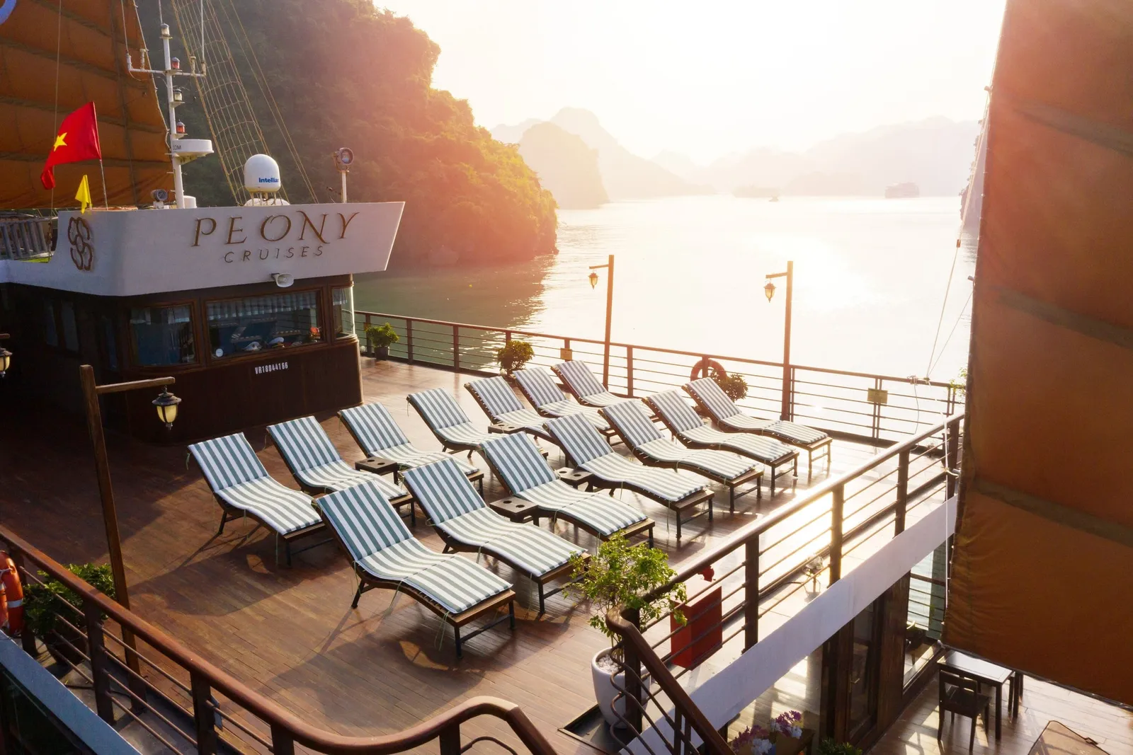 A wooden sun deck with rows of green-and-white striped loungers on a junk-style boat, with a Vietnamese flag, orange sails, and a 'Peony Cruises' sign on the cabin, looking out over calm water and limestone karsts at sunset.