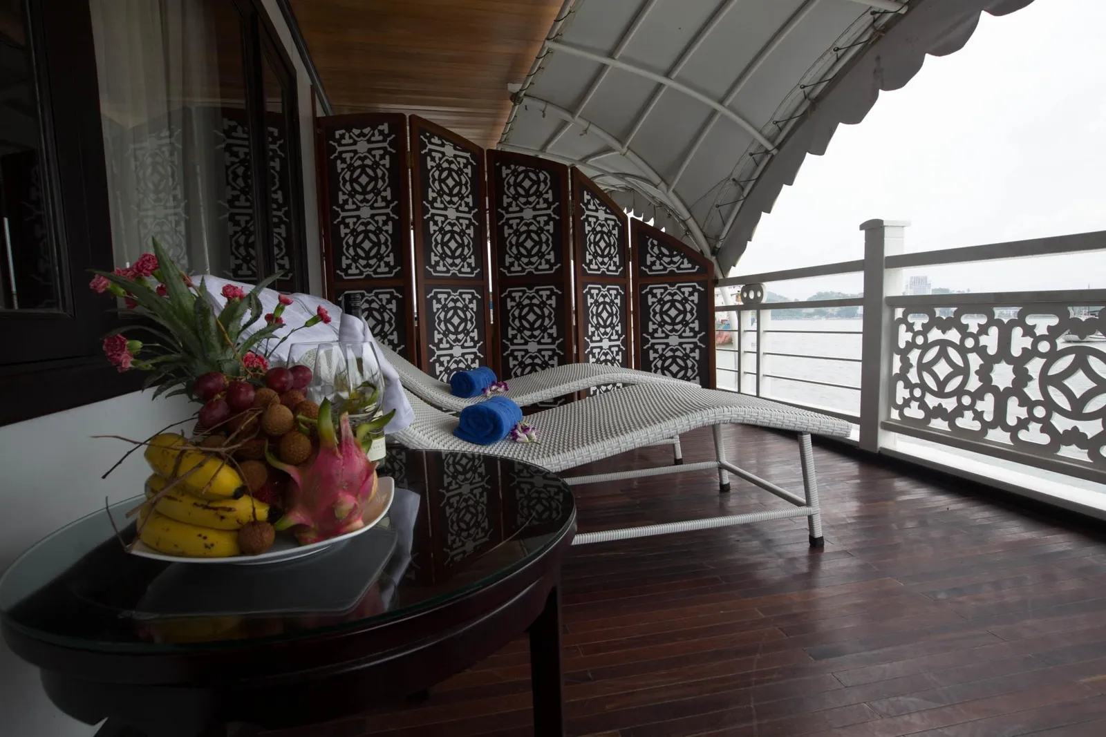 A private balcony deck with dark wood flooring, white wicker loungers with rolled blue towels, a carved wooden screen, and a glass table with a fruit plate, overlooking calm water under a gray sky.