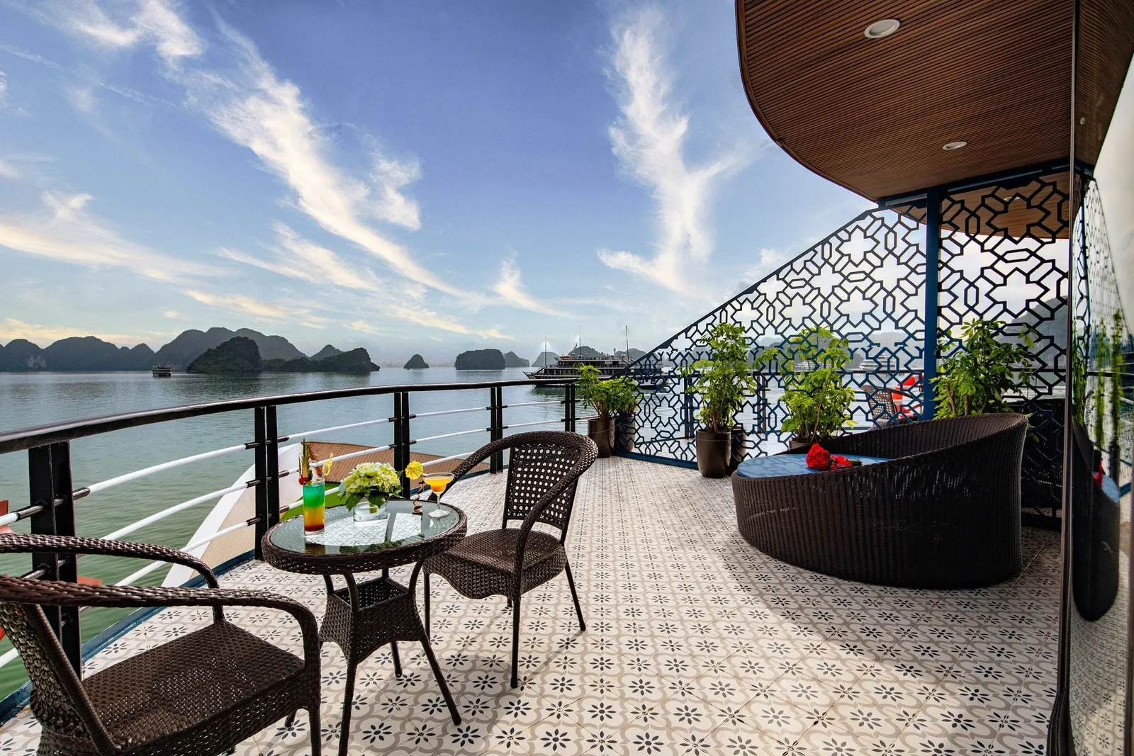 A tiled cruise-ship deck with wicker chairs, a small glass table with drinks and flowers, a daybed and potted plants beside a lattice screen, overlooking calm water and limestone karsts with another boat in the distance.