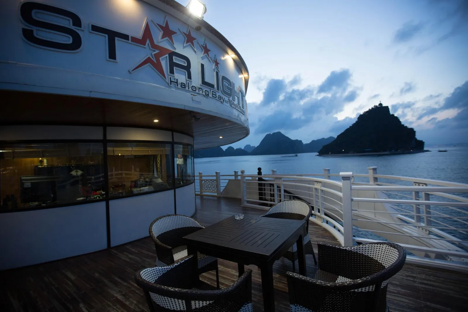 A wooden deck at dusk with a dark table and wicker chairs beside a white railing, with the 'Starlight Halong Bay' name and red stars on the curved cabin wall and limestone islands over calm water in the background.